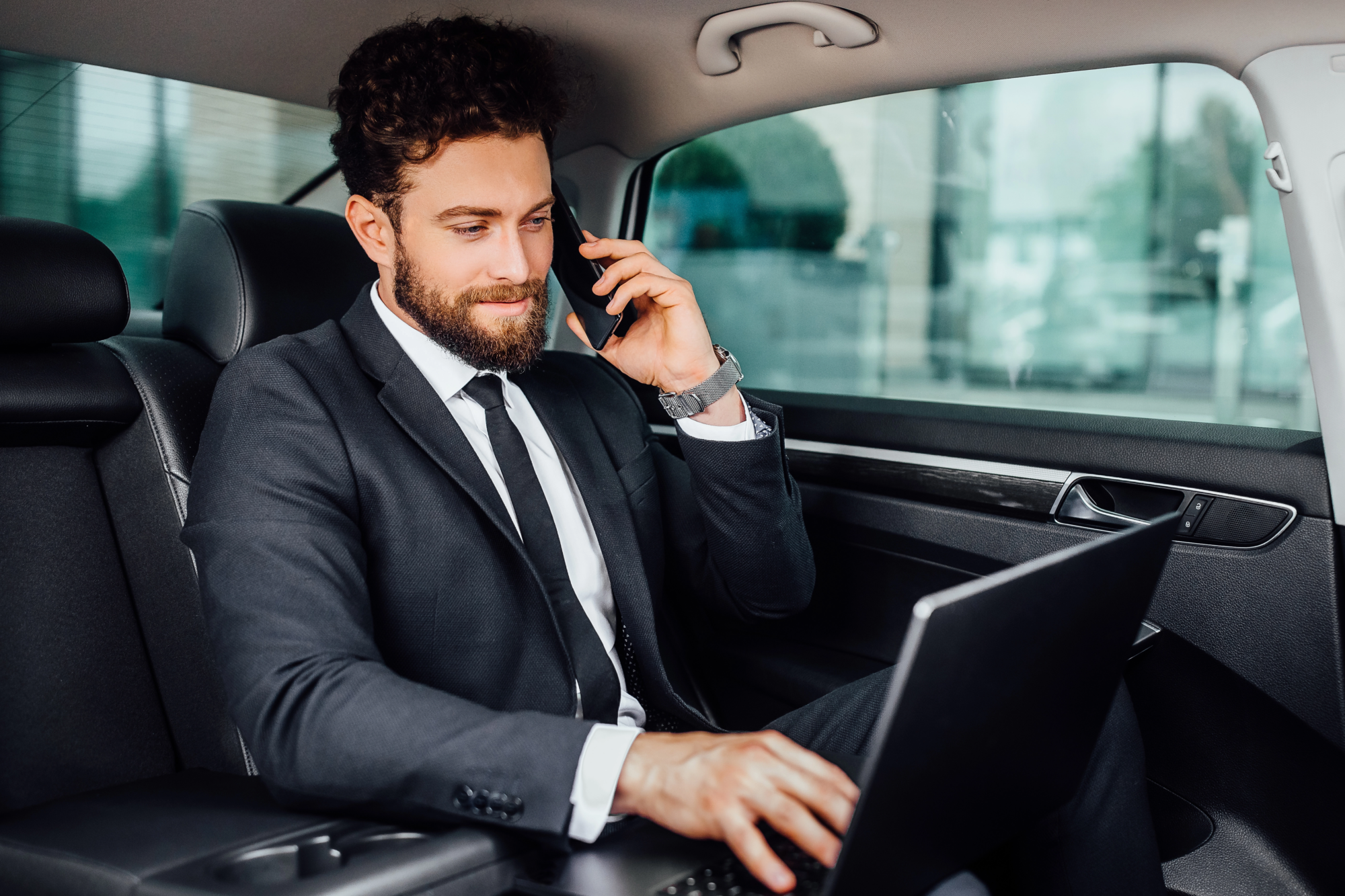 handsome-bearded-smiling-businessman-working-his-laptopand-speaking-mobile-phone-backseat-car